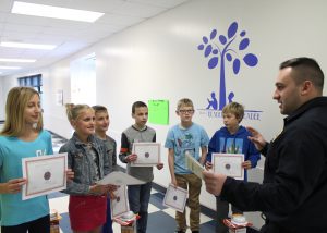 Firefighter stands in school hallway with group of students handing them award certificates.