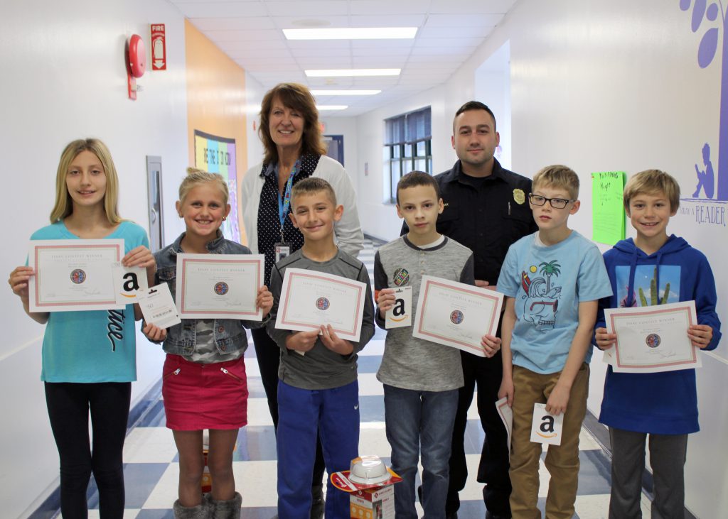 A group of students pose holding their award certificates. They are joined by their school principal and the firefighter who presented the awards.