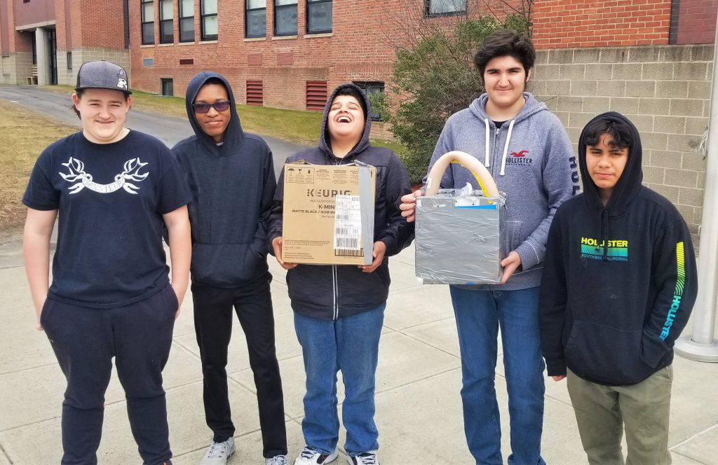 Standing outside a school building, five students pose for a group photo. Two are holding an engineering project built inside carboard boxes.
