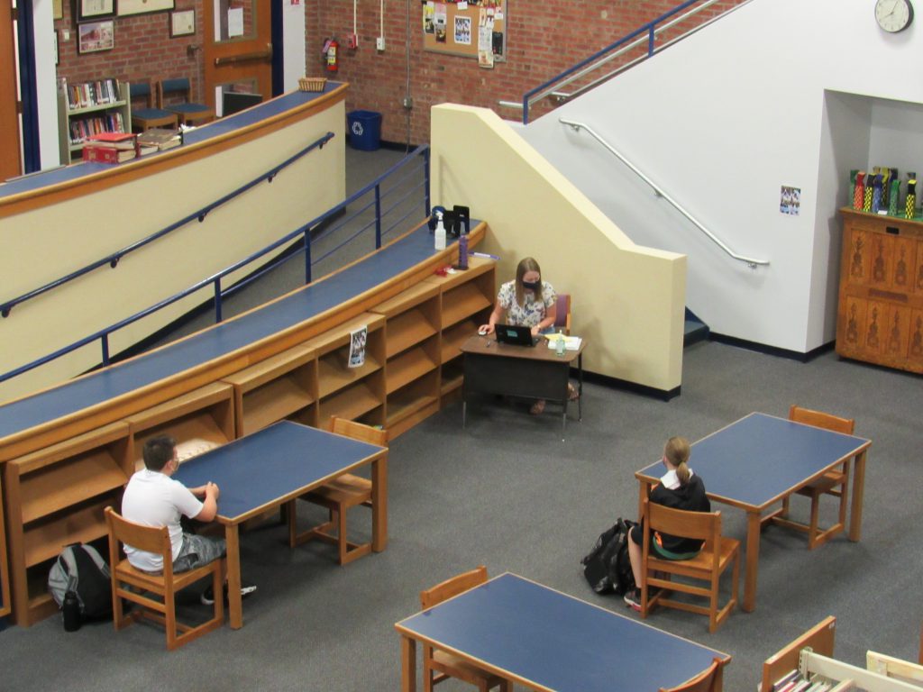 looking down from a balcony, there is a teacher at the front of the room and a couple of students sitting at desks far apart.