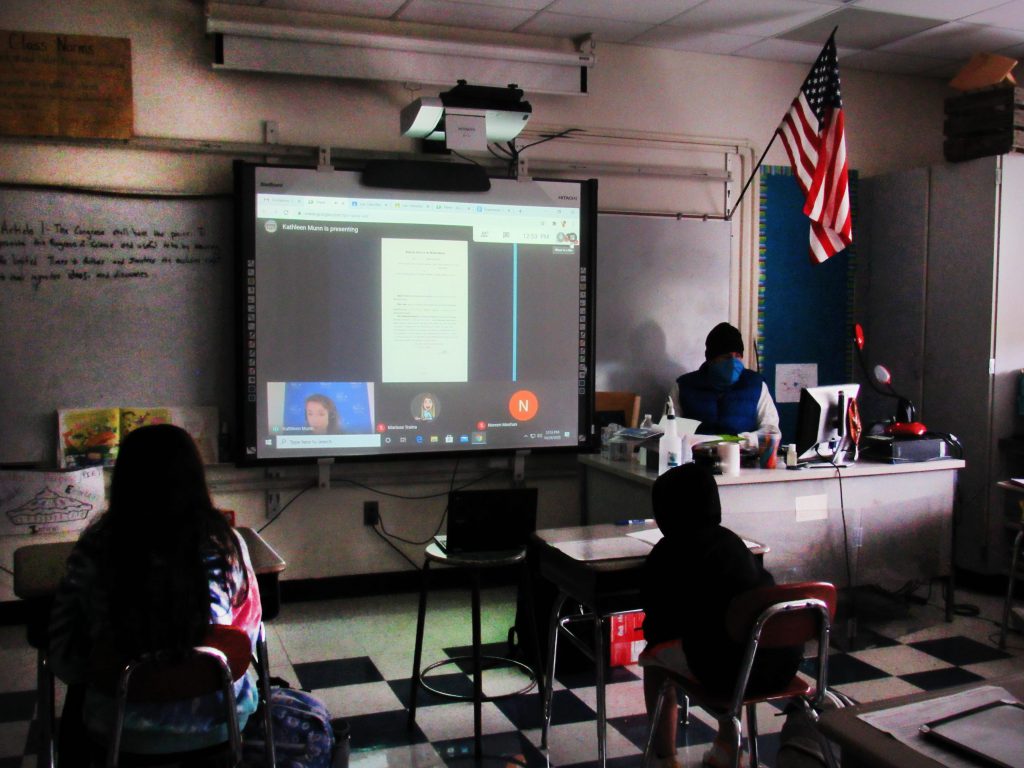 Students sit at desks and watch a screen with a live interactive guide