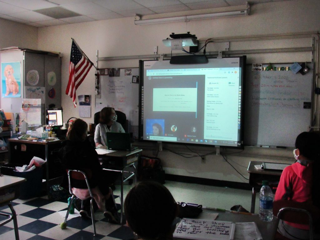 Students sitting at desks watch a screen with information about the National Archives and a live person in the corner talking to them.
