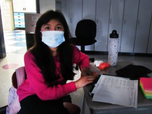 A girl with long dark hair wearing a pink sweater and a face mask watches the screen for the program. 