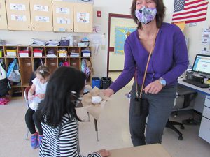 A woman holds a snowball in her hand on a piece of brown paper towel showing it to a kindergarten student. The woman is laughing behidn her mask.