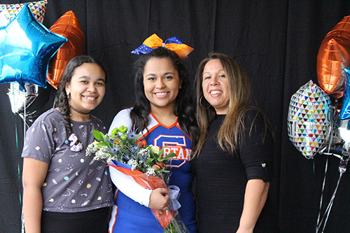 Three women smiling. The one in the center is a young woman in a cheerleader uniform wearing blue and orange bow in her hair, holding flowers. Balloons are on the left sie of them.