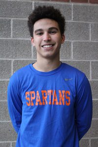 A young man with dark hair and a big smile wears a blue longsleeve t-shirt that says Spartans in orange on it. background is a brick wall.