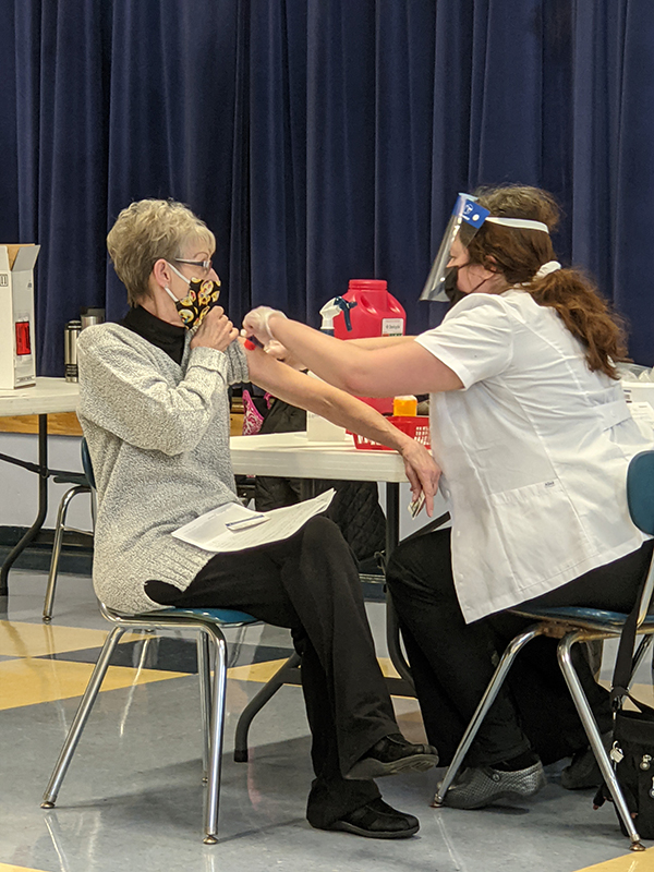 A woman with short light hair wearing a mask pulls up her left sleeve while another woman with a mask and face shield gives her a vaccination.