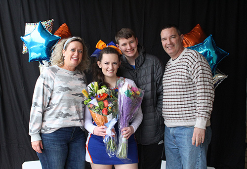 Four people standing in front of blue and orange balloons. A woman on the left wit blonde hair and a light colored sweater, a young woman in a blue and orange cheerleading uniform, holding two bunches of flowers and wearing an range bow in her hair, two men on the right. All are smiling.