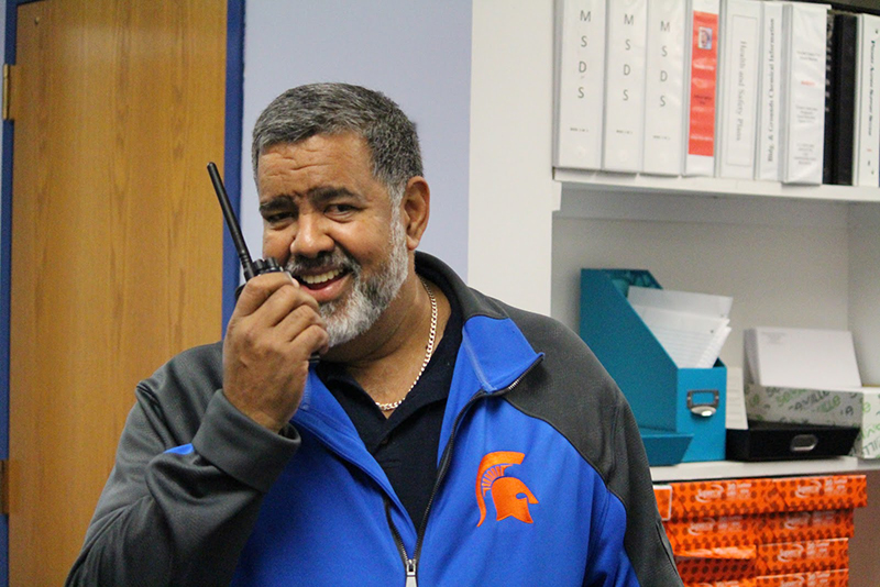 A man with salt and pepper hair and beard smiling. He is holding a walkie-talkie up to his mouth. He is wearing a blue jacket with an orange spartan logo on the front right chest.