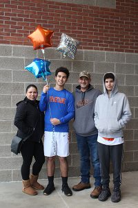 A family of four people, a mom and dad and two sons. One is holding balloons. All are smiling.