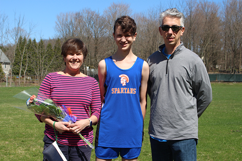 A woman on the left and a man on the right with a young man in the middle. The young man is wearing a blue runners shirt with a gold spartan. The woman is holding flowers.