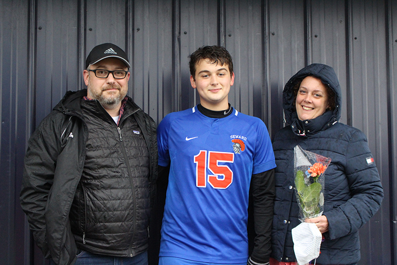 A high school senior boy with short dark hair wearing a blue soccer jersey with number 15 in orange. He is flanked by a man wering glasses, baseball cap and black vest and a woman wearing a winter jacket with hood holding pink flowers.