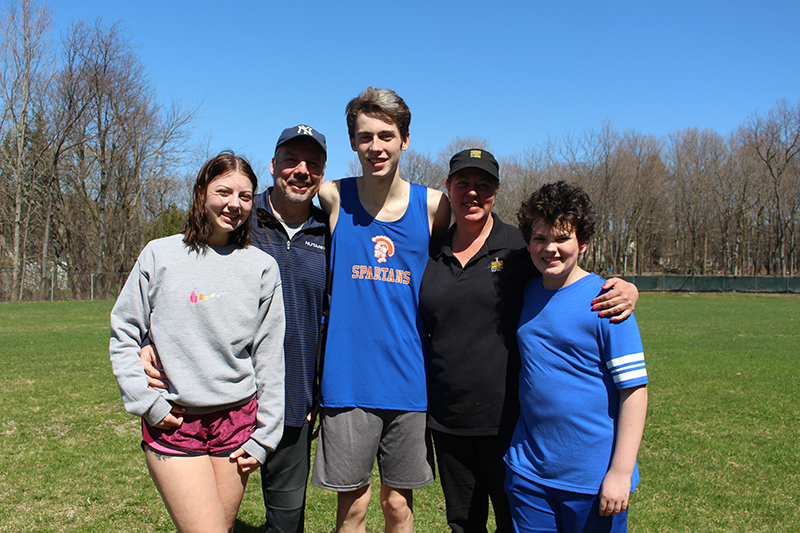 A young man in the center of five people. He has blone hair and is wearing a blue athletic shirt with a gold spartan on it. On either size are two other people a girl, man, woman and boy.