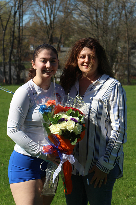 A high school age young woman with dark hair in a ponytail holds a bouquet of flowers. She is wearing a long sleve white shirt an blue shorts. She is standing next to a woman with long dark hair ane a light blue shirt