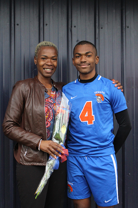 A high school senior boy with short dark hair wearing a blue soccer jersey with an orange number four. He is flanked by a woman in a brown jacket and flowered shirt holding flowers. They are both smiling.