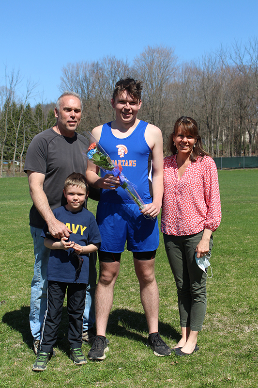 A tall young man stands with a man on his left and a woman on his right. A little boy stands in front of the man. The young man wears athletic shorts and a blue athletic shirt with a spartan on it.