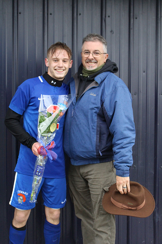 A high school senior boy with short light hair wearing a blue soccer jersey has his arm around a man wearing glasses and a blue jacket with a hood. Both are smiling.