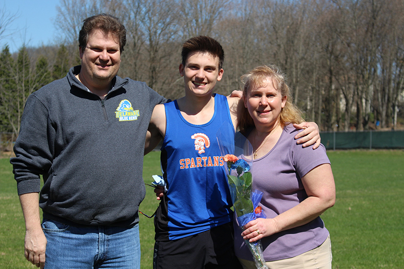 A young many with dark hair is in the center wearing a sleevless athletic shirt, blue with a gold spartan on it. On the left is a man and on the right is a woman holding a flower. All are smiling.