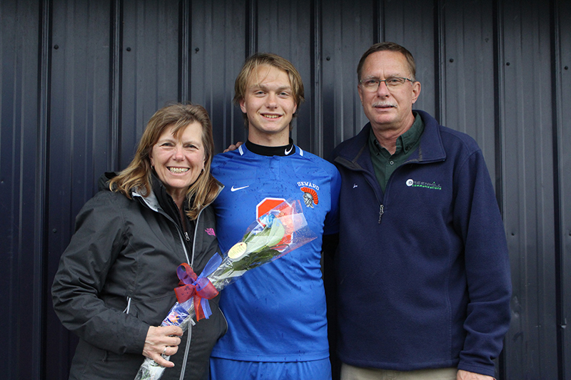 A high school senior boy wearing a blue soccer jersey with an orange number 3 has an arm around a woman smiling holding flowers and a man wearing a blue jacket.