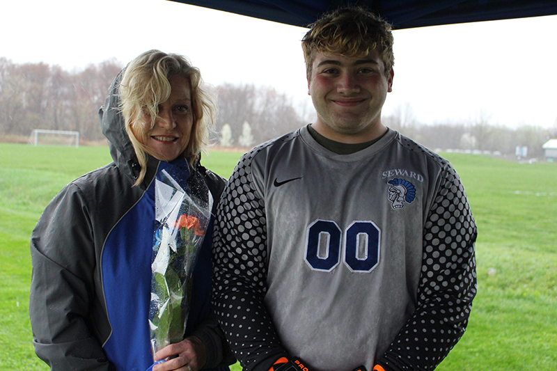 A high school senior boy waring a gray soccer jersey with double zeros in blue smiles. Next to him is a woman with blonde hair wearing a jacket with hood and holding flowers. They are under a tent, it looks like its raining.