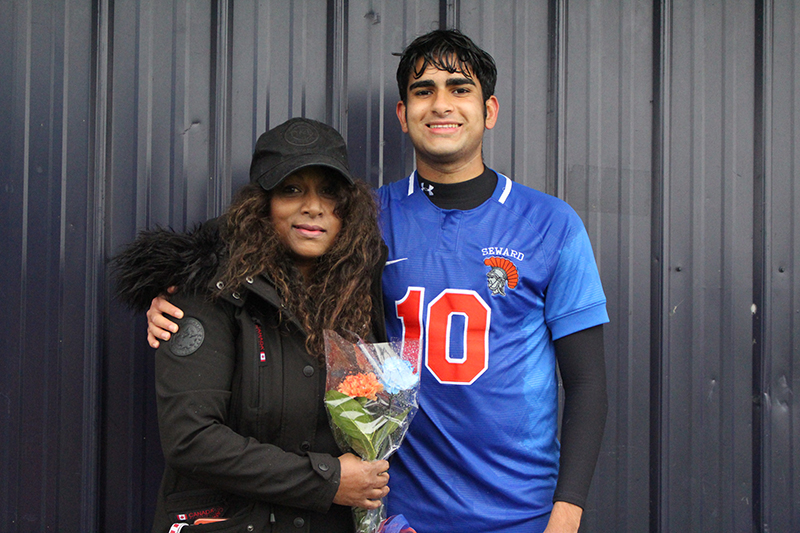 A high school senior boy wearing a blue soccer jersey with number 10 in orange smiles and has his arm around a woman wearing a winter jacket and hat, holding flowers.