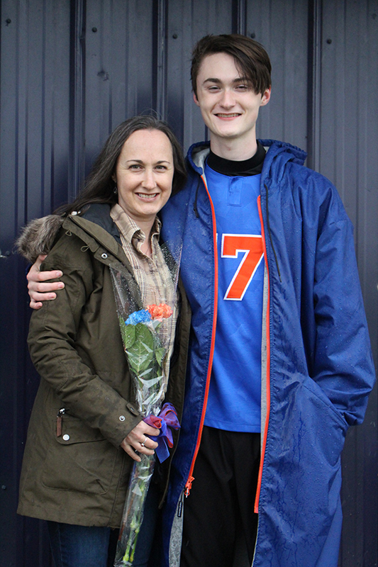 A high school senior boy wearing a blue soccer jersey with an orange number 7 on it smiles. He has dark hair and is wearing a  blue winter jacket. He has his arm around a woman with long brown hair, wearing a green winter jacket. They are smiling.