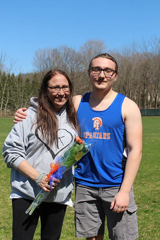 A young man with short dark har and wearing glasses stands with his arm around a woman with long hair holding a flower. The young man is wearing a blue sleeveless athletic shirt with a gold spartan on it. 
