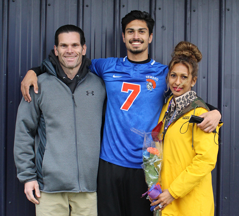A high school senior boy wearing a blue soccer jersey and the number 7 in orange smiles broadley. He has dark hair and has an arm around a man with dark hair and green jacket and a woman with brown hair piled on top of her head, wearing a yellow jacket holding flowers. 