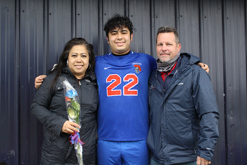 A high school senior soccer player wearing a blue soccer jersey with the number 22 in orange on it. He has dark hair and has his arm around a woman with dark hair wearin ga winter jacket holding a flower and a man with darker hair wearing a blue winter jacket. All are smiling.