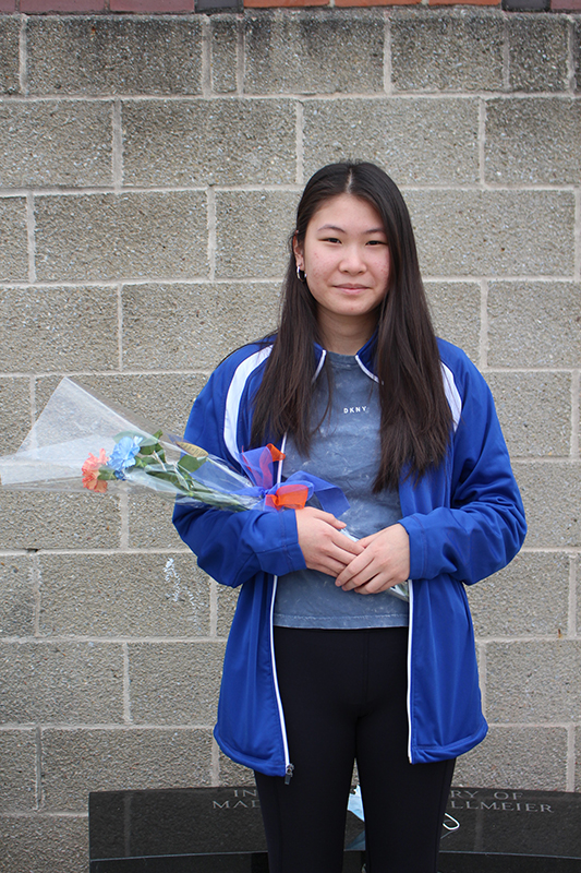 A young woman with long dark hair dressed in blue holds a flower.