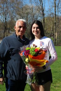 A high school age young woman is on the right, she has long dark hair wearing a long sleeve white shirt and blue shorts. She is holding multi-colored flowers. On the left is a man with gray hair and wearing a blue jacket. They are both smiling.