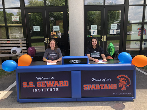 A large table with Ss Seward spartans written on the front. On either side is a young woman. On the left is a young woman wearing a gray tshirt that says Eastern Warriors. she has long light brown hair and there is a paper in front of her. She is smiling. On the right is a young woman with long dark hair. She is wearing a black tshirt with Morrisville Basketball written on it. She is smiling too. There are balloons all around.