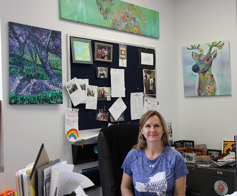 A woman with shoulder-length blonde hair sits at a desk smiling. She is wearing a light purple shirt with the state of New York on it . Inside the graphic it says Seward Spartans. Surrounding her on th ewalls is all artwork done by students.