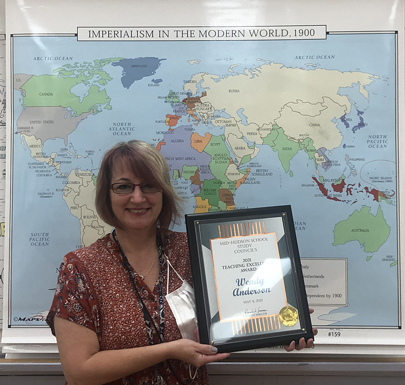 A large map of the world is the background. In front is a woman with chin-length blond hair holding an award. It's a certificate in a black frame..