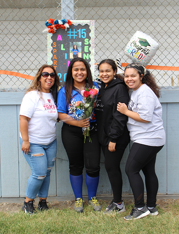 Four women all smiling. The one that is second from left is wearing a softball uniform and holding flowers. There are balloons in the background. All are smiling.