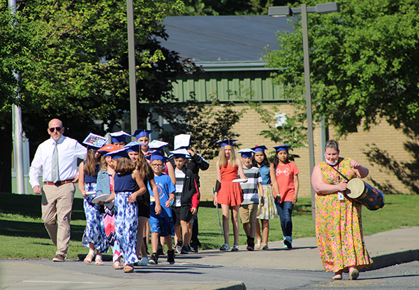 A line of fifth-grade students, all wearng graduation caps, are walking toward the camera. On the right is a teacher wearing a long yellow dress and playing a drum.