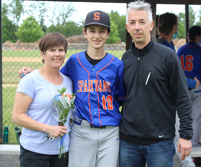 A young man in the center wearing a baseball uniform - a blue shirt with Spartans written in orange and a black baseall cap with an S on it. Next to him is a woman holding flowers on the left. She has short brown hair. On the right is a man in a black jacket with gray hair.