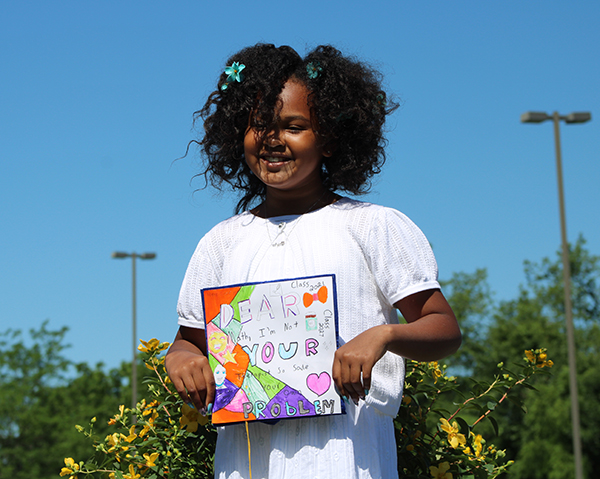 A girl with a big smile and wearing a white shirt holds her decorated graduation cap in front of her. She is smiling and has chin-length curl black hair.