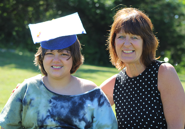 A woman with a black and white polka dot dress smiles as she poses with a fifth-grade student. The student is a young woman with a tie-dye shirt. She is wearing a blue graduation cap. They both have short brown hair and are smiling.