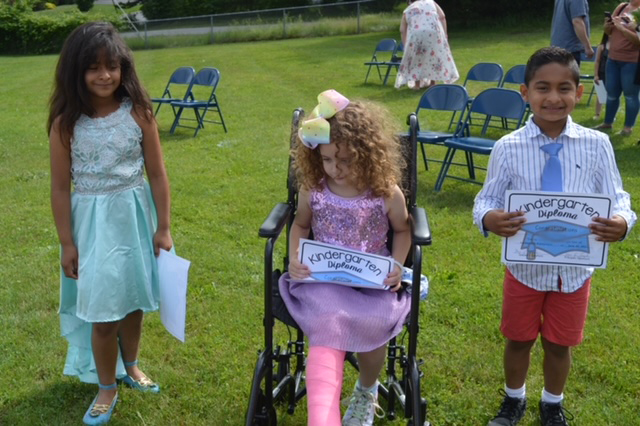 Three kindergarten children, a girl on the left with long dark hair and a light green dress, a girl in the center with a pink cast on her leg, a purple dress and a yellow bow in her long curly hair, and a boy on the right, wearing red shorts and a white shirt. All are holding kindergarten certificates.