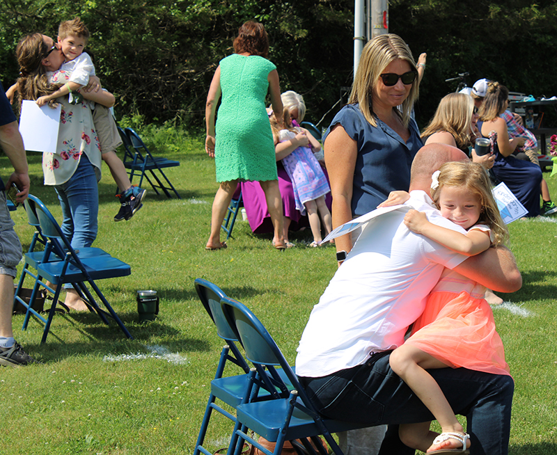 A little girl hugs her dad who is sitting in a chair. She has blonde hair and is wearing an orange dress.