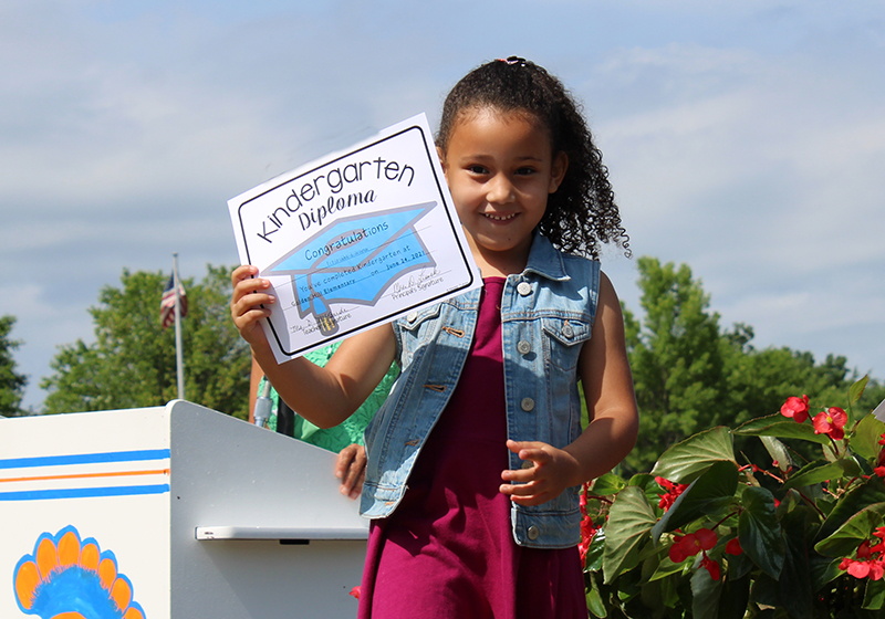 A kindergarten girl holds up her kindergarten diploma. She is wearing apink dress and a enim jacket. She has long curly dark hair that is pulled back. In the background is a blue sky and green trees.