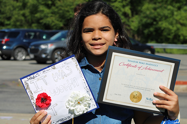 A fifth-grade girl with long dark hair smiles and holds up two certificates she received.