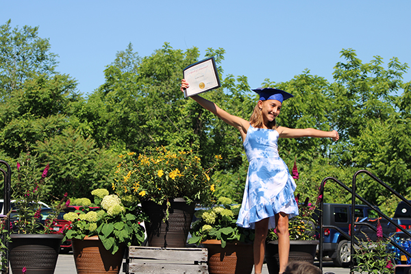 A fifth grade girl wearing a blue graduation cap and a blue and white dress. holds her certificate in her right hand way up high and her left arm is stretched out on the other side of her. Behind her is blue sky and green trees. 