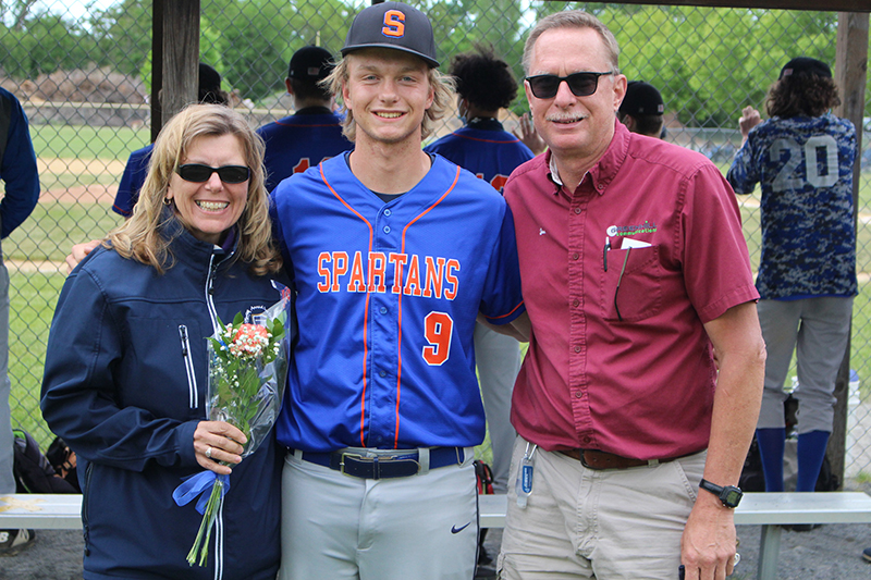 A tall young man with longish blonde hair is in the center. He is wearing a baseball uniform - blue shirt with Spartans written in orange, gray pants and a black cap with the letter S on it. On his left is a woman with shoulder-length blonde hair, wearing a blue zipped jacket and holding flowers. On his right is a man with short brown hair, gray mustache, wearing sun glasses, a red shirt and light pants. They are smiling.