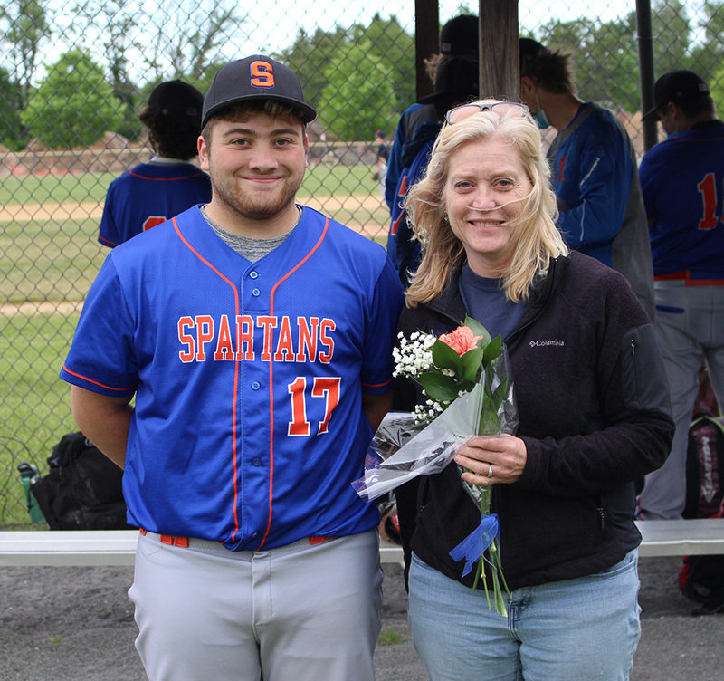 A young man wearing a baseball uniform - blue shirt with Spartans written in orange and gray pants - stands with a woman with blonde hair olding flowers. They are both smiling.