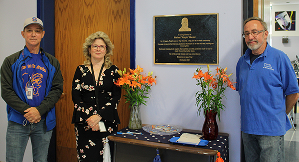 A bronze plaque hands on a wall. There are orange lilies on a table on either side of the plaque. Three people stand near it, a man and  woman on the left, and a man on the right. 