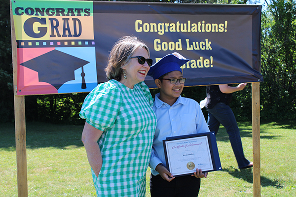 A teacher wearing a green check dress stands with a young male student who is holding his certificate. He has on a blue button-down shirt, glasses  and a blue graduation cap. They are in front of a sign that says Congratulations! Good Luck 5th Grade! 