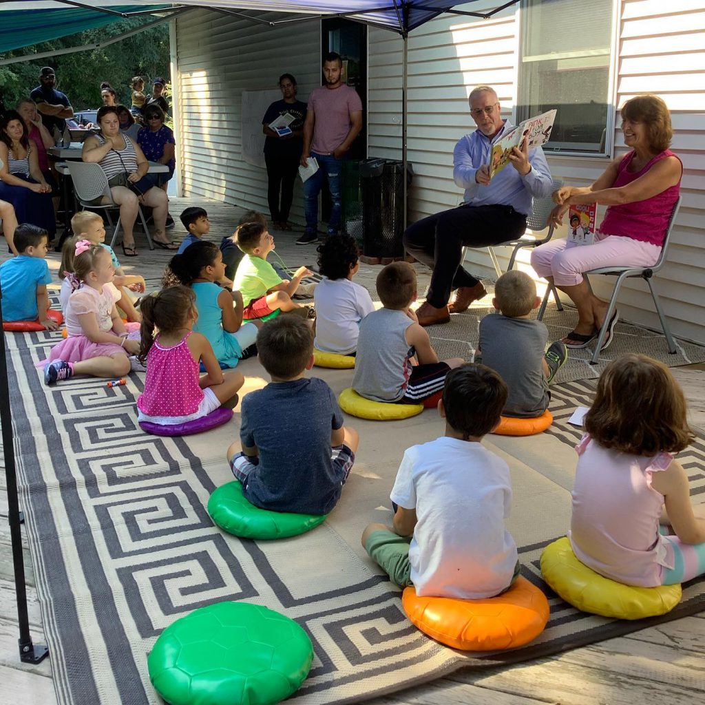 A group of about a dozen kindergarten students sit on colorful round pillows outdoors. Two adults, a man and a woman, sit in front of them. The man is holding up a book while he reads as the woman smiles.