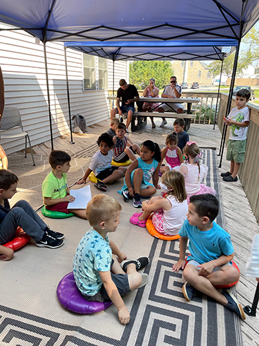 A group of about a dozen kindergarten students sit on colorful pillows outside the library, with parents in the background.